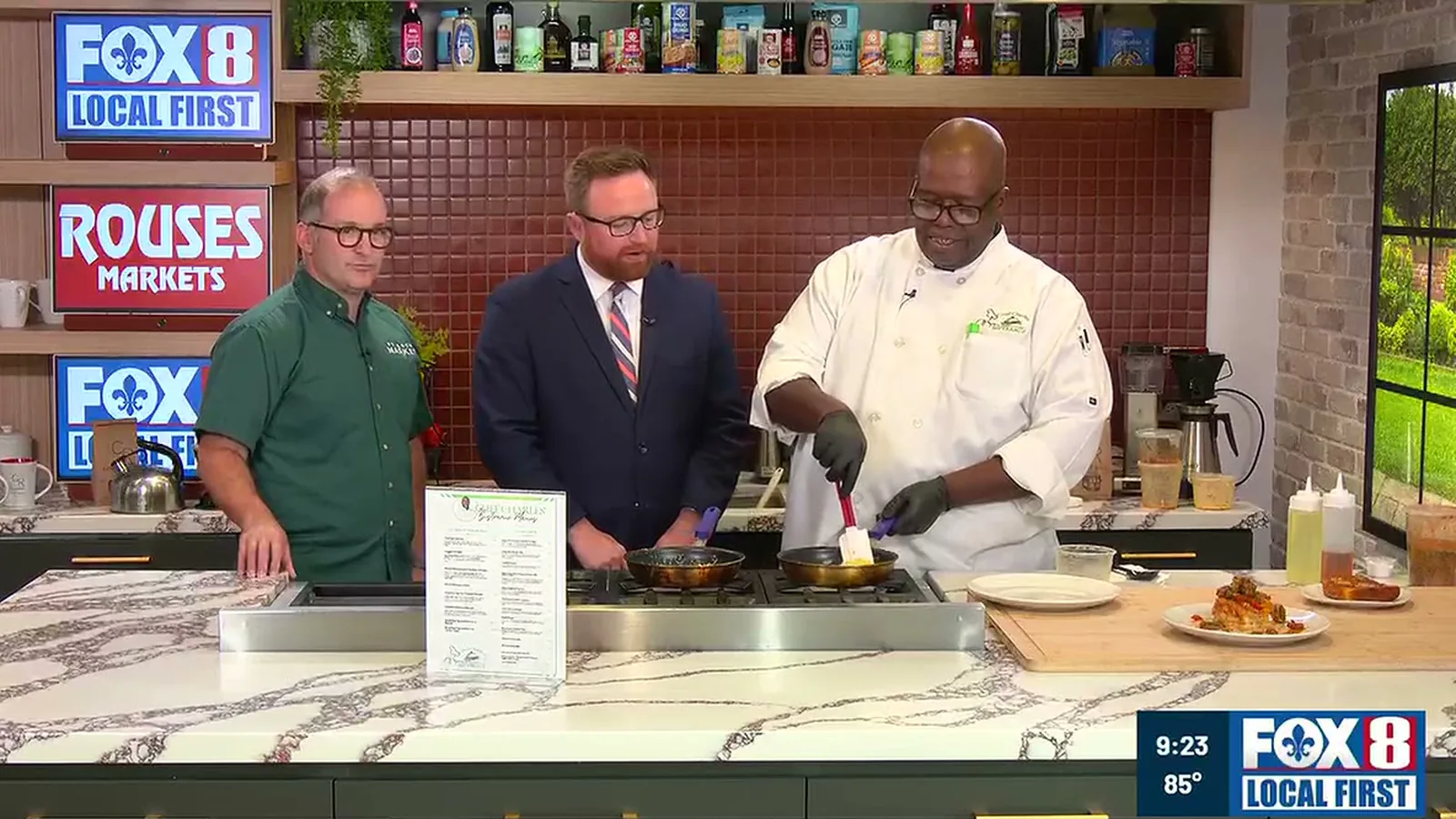 Kevin Pedeaux, host Rob Krieger, and Chef Charles Wilford Sr. at the stove in the FOX 8 studio kitchen, with two pans cooking and plated dishes on the counter