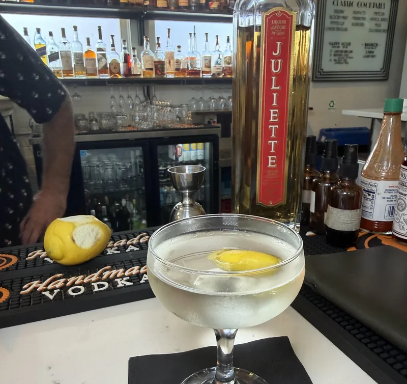 A bottle of Juliette peach liqueur with its red label standing on the bar next to the finished cocktail in a coupe, a cut lemon, and a jigger