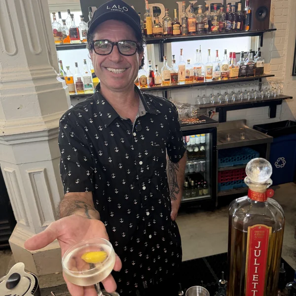Bartender Julien Freed holding out the Juliette cocktail in a chilled coupe at Roch's Reprieve, with the Juliette bottle beside him