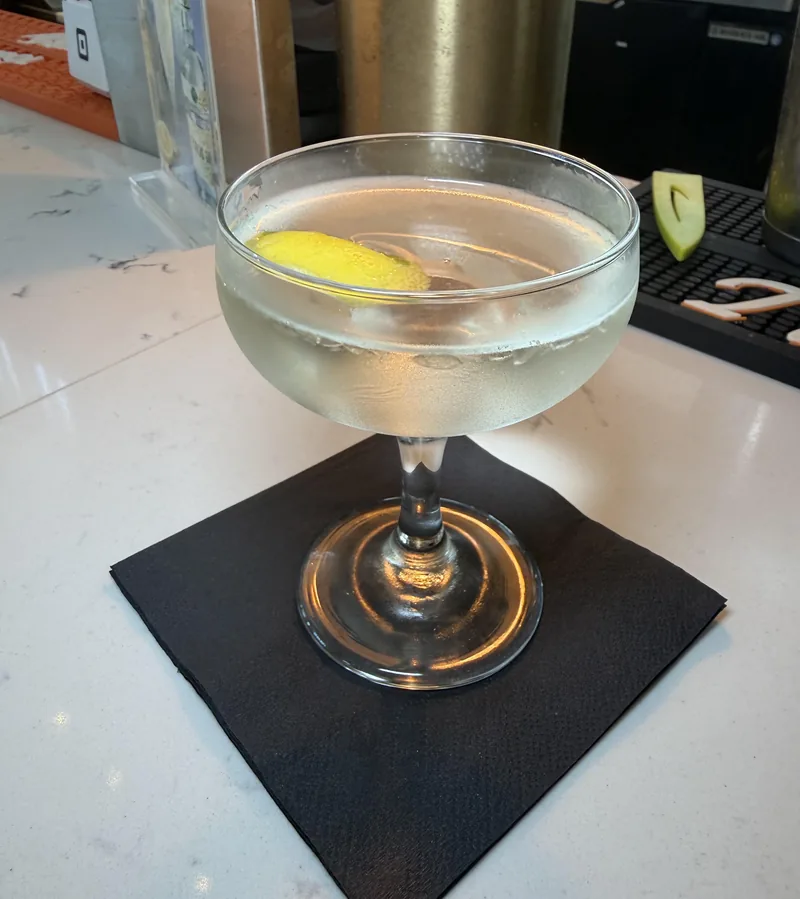 The finished Juliette cocktail, pale gold and cloudy with cold, in a chilled coupe on a black napkin with a lemon twist floating on top