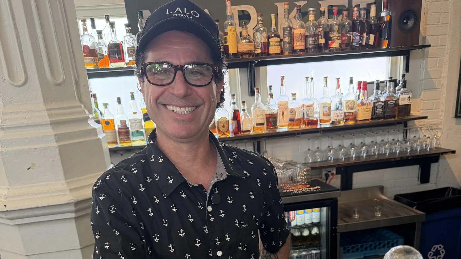 Bartender Julien Freed smiling behind the bar at Roch's Reprieve in a black anchor-print shirt and a ball cap, with the backbar bottles behind him