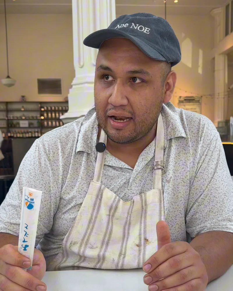 Alex Regules in a cap and apron talking at the counter of St. Roch Market, holding a chopsticks sleeve