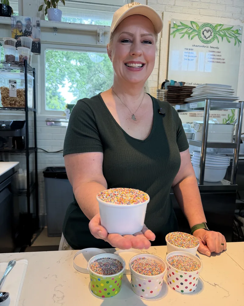 Alison Grice Barrios smiles behind the Olive & Co counter at St. Roch Market, holding out a sprinkle-covered dot cake with more dot cakes on the counter below